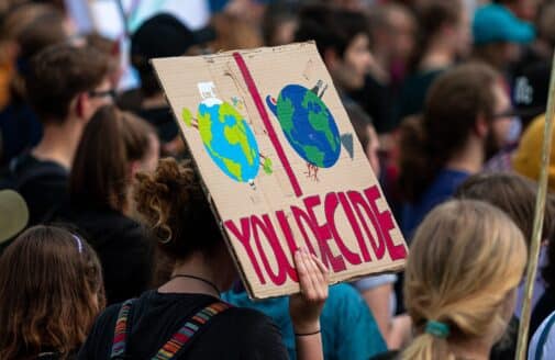 a hand emerges from a crowd of people facing away from the camera, holding a protest poster painted on cardboard. the poster depicts two planet earths, one with trees and the other with natural disasters, and includes the text YOU DECIDE